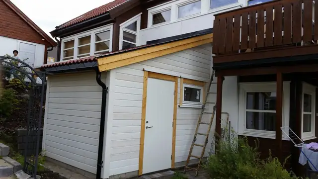 Boden sett fra bakkenivå. Veggen har hvit, liggende kledning, en hvit dør og et lite vindu med umalte lister rundt. På hjørnet går et svart nedløp fra takrenna, og en stige står inntil veggen.