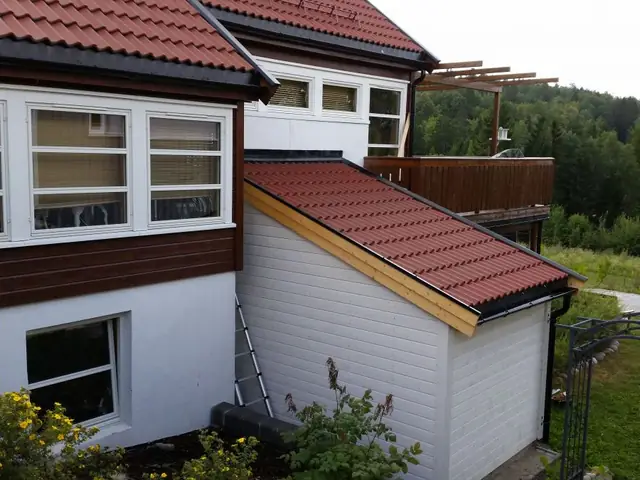Hvit bod med skrått tak med rød takstein, bygget inntil et hus i hvitt og brunt. Bildet er tatt ovenfra, med plen og skog i bakgrunnen.