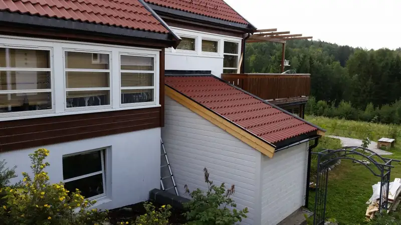 Hvit bod med skrått tak med rød takstein, bygget inntil et hus i hvitt og brunt. Bildet er tatt ovenfra, med plen og skog i bakgrunnen.