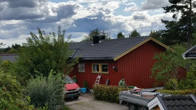 Huset sett ovenfra mot gavlen. Gavlveggen er rød med gule vindskier, og taket er dekket med svart takstein. På gårdsplassen står en rød bil og en tilhenger, og en mann står ved veggen.