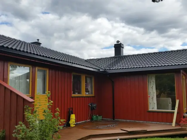 Rødt hus i én etasje med gule vinduskarmer, sett fra hagen med terrassen foran. Taket er dekket med svart takstein, og pipa, takrenna og nedløpet er svarte.