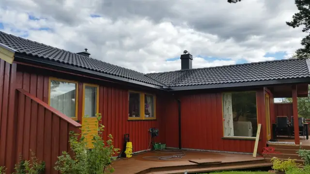 Rødt hus i én etasje med gule vinduskarmer, sett fra hagen med terrassen foran. Taket er dekket med svart takstein, og pipa, takrenna og nedløpet er svarte.