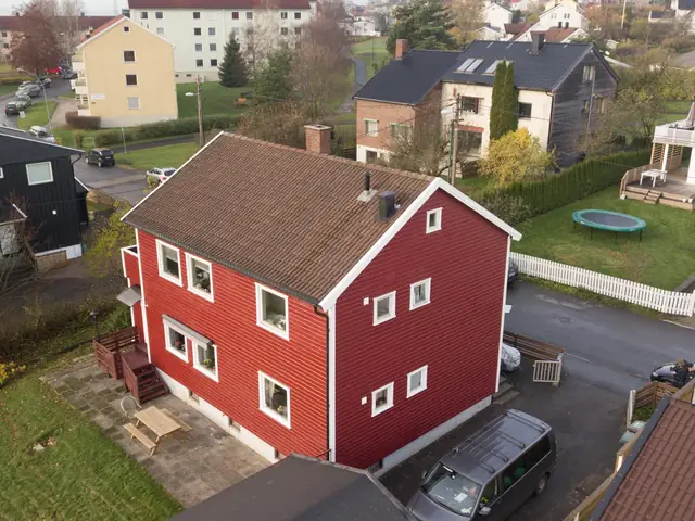 Dronebilde av rødt hus i to etasjer med liggende panel, hvite vindusomramminger og brun takstein. Nabohus og hager ligger rundt.