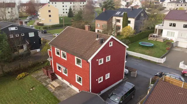 Dronebilde av rødt hus i to etasjer med liggende panel, hvite vindusomramminger og brun takstein. Nabohus og hager ligger rundt.