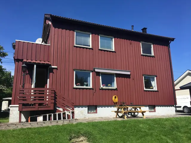 Mørkerødt hus med stående kledning, fotografert fra plenen. Til venstre et utbygg med dør og balkong over, foran veggen et bord med benker.