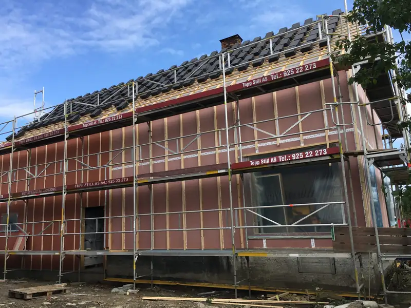 Hus under arbeid med stillas langs veggen. Veggen er dekket av rosa vindsperre med lekter, og det ligger mørk takstein på en del av taket.