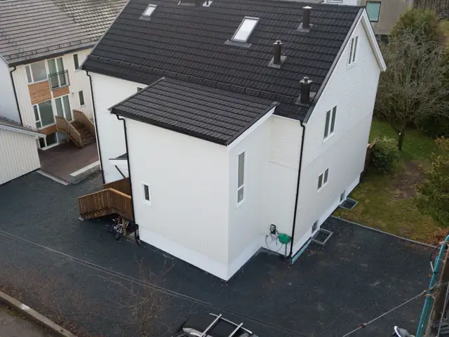Dronebilde av et lyst trehus med svart tak. Midt i bildet står tilbygget i to etasjer med stående kledning og eget tak, og til venstre for det er det en utetrapp i tre.
