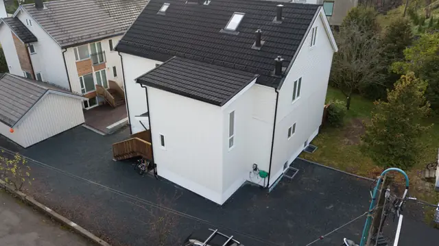 Dronebilde av et lyst trehus med svart tak. Midt i bildet står tilbygget i to etasjer med stående kledning og eget tak, og til venstre for det er det en utetrapp i tre.