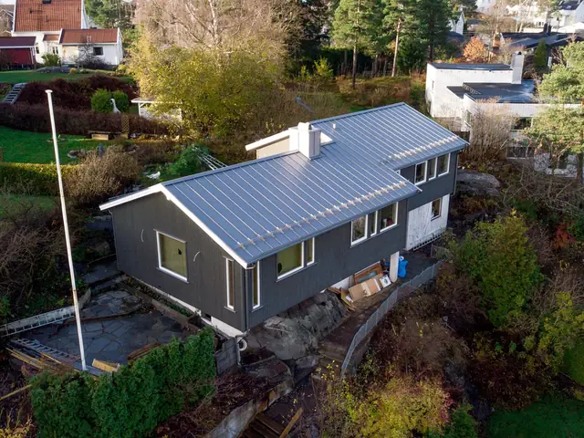Dronebilde av et hus i skrånende terreng med mørkegrå, stående kledning, hvite vinduer og grått metalltak. Nedenfor huset ligger papp og sekker, og stiger ligger i hagen.