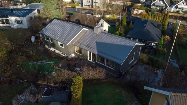 Dronebilde ovenfra av huset med grått metalltak og mørkegrå kledning. Langsiden har en høy glassdør og to store vinduer. Foran ligger en platting av tre, og to personer står ved hjørnet.