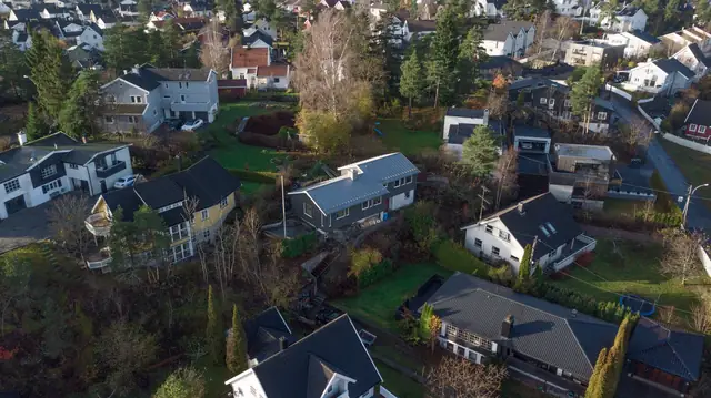 Dronebilde av et boligområde med hager og hus. Midt i bildet ligger huset med mørkegrå kledning og grått metalltak.