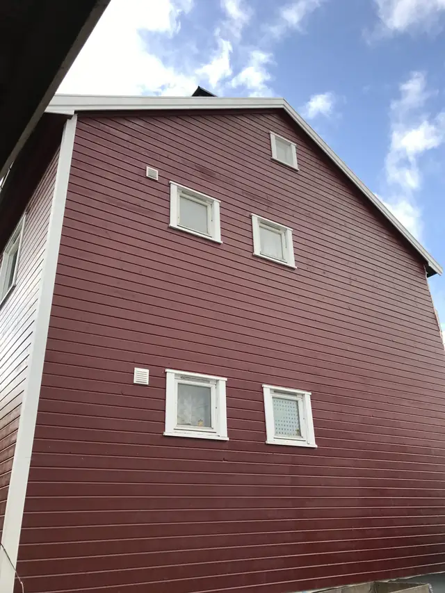 Rød gavlvegg fotografert nedenfra mot himmelen. Fem vinduer med hvite omramminger og to hvite ventiler i veggen.