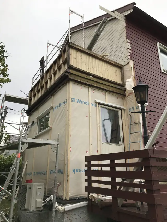 Utbygg dekket med lys vindsperreduk og stående lekter, med stillas inntil. Gavlveggen bak balkongen har lyst, liggende panel, og veggen til høyre har mørkerødt, liggende panel.