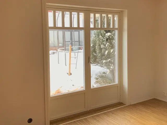 Hvit tofløyet dør med store glassfelt og en rad smale ruter øverst, i et tomt rom med lyst tregulv. Utenfor ligger det snø i hagen.