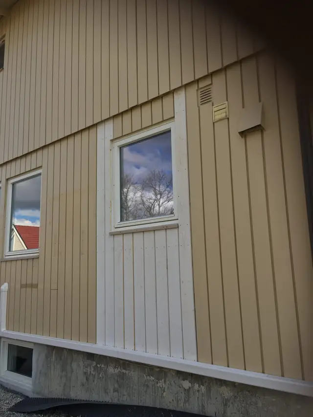 Yttervegg med beige stående kledning, sett på skrå nedenfra. På begge sider av og under et hvitt vindu er kledningsbordene hvite i stedet for beige, og nederst ses grunnmuren.