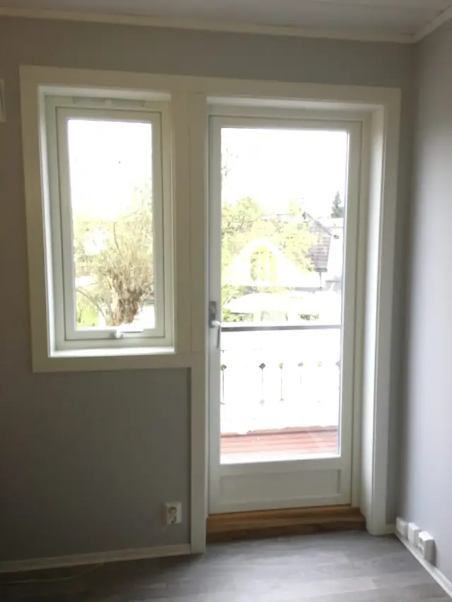 Rom med grå vegger der et smalt hvitt vindu og en hvit balkongdør med glass står ved siden av hverandre med hvite lister rundt. Utenfor ses en balkong med hvitt rekkverk.