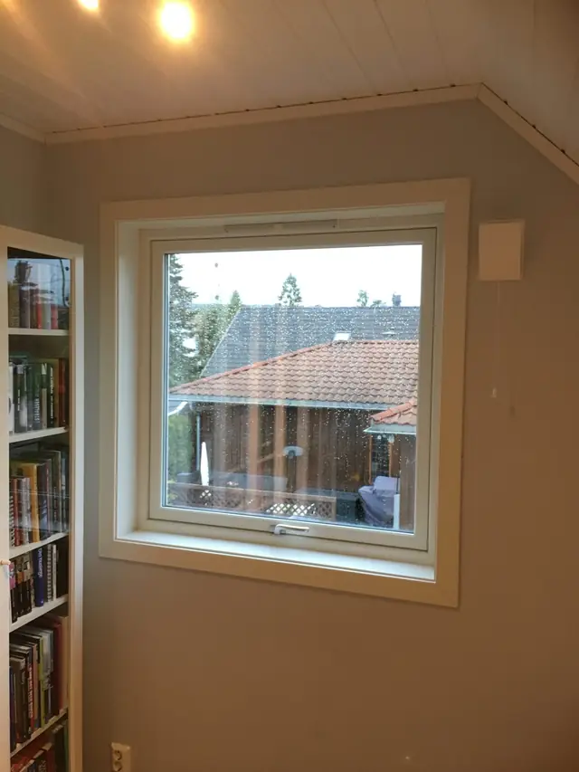 Kvadratisk hvitt vindu med én ramme i en gråblå vegg under skråtak med hvitt panel. Det er regndråper på glasset, og utenfor ses tak med rød takstein.