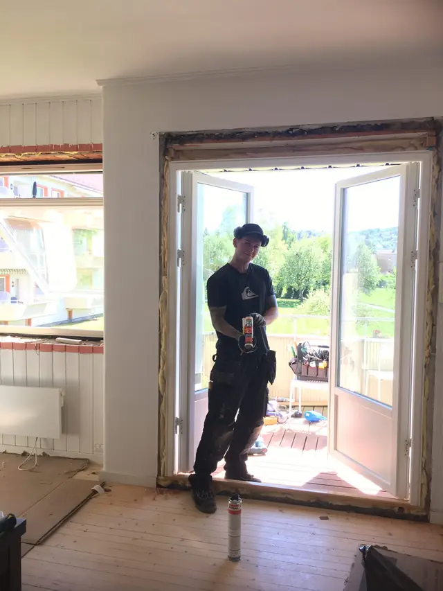 Rom sett innenfra med en hvit tofløyet balkongdør som står åpen. Veggen rundt karmen er åpen med synlig isolasjon. En håndverker står i døråpningen, og verktøy ligger på balkongen.