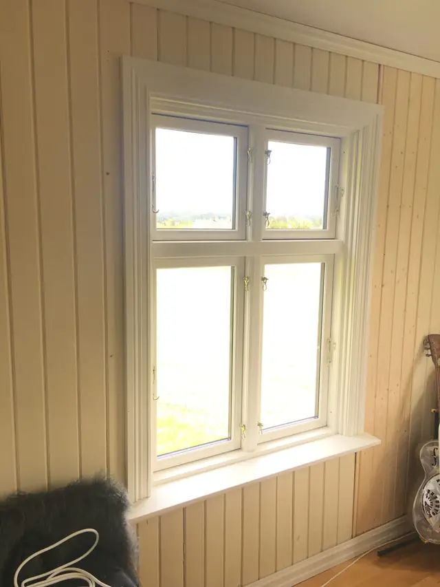 Hvitt vindu med to små rammer øverst og to høye nederst, sett innenfra i et rom med lyst stående panel på veggene. Gjennom glasset ses et åpent landskap.