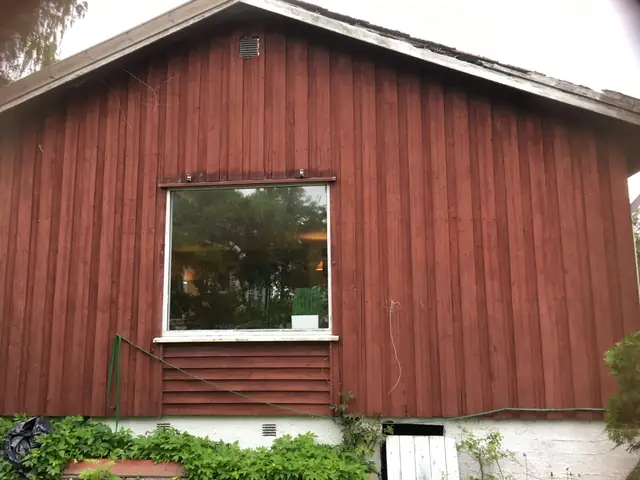 Gavlvegg med rød, stående kledning, ett stort vindu med et felt av liggende panel under, og værslitte vindskier øverst.