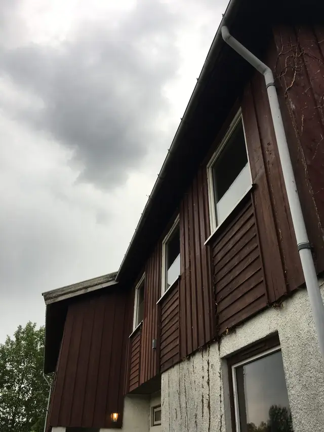 Langvegg sett nedenfra: mørk rødbrun kledning, tre vinduer med felt av liggende panel under, hvitpusset mur med et vindu nederst og et hvitt nedløpsrør.