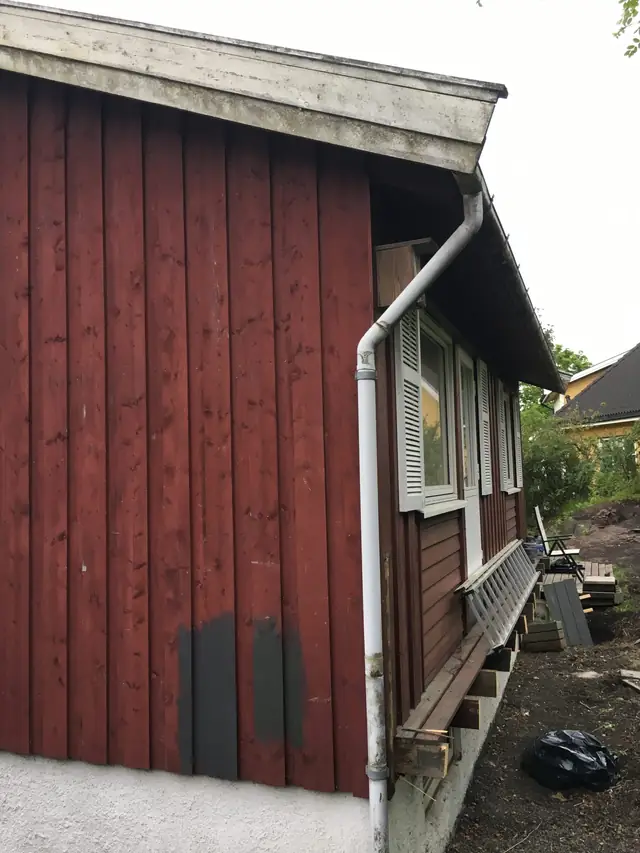 Hushjørne med rød, stående kledning der to felt nederst er malt grå. Langs den andre veggen er det vinduer med grå skodder, og en stige ligger på bjelker som stikker ut fra veggen.