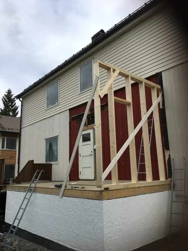 En vegg av nye trestendere står på et gulv over en hvit, pusset mur, støttet av skråstilte bord. Bak er husveggen med rød kledning og en hvit dør.