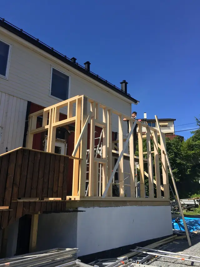 Reisverk av lyse trestendere på en hvit, pusset mur inntil huset. En håndverker står på et stillas inne i reisverket, og til venstre er det et brunt rekkverk i tre.