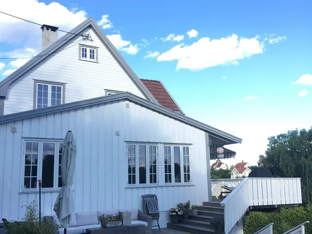 Hvitt trehus med grå vindskibord og smårutede vinduer under blå himmel. Foran den lave fløyen ligger en terrasse med hagemøbler, og til høyre en trapp med hvitt rekkverk.