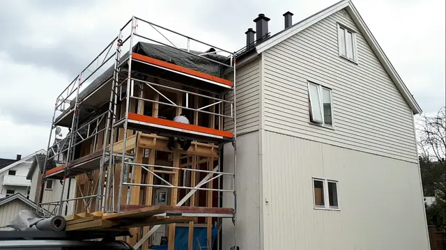 Tilbygget som åpent reisverk i to etasjer, omgitt av stillas og dekket til på toppen. Til høyre er gavlveggen på huset, med liggende kledning øverst og stående nederst.
