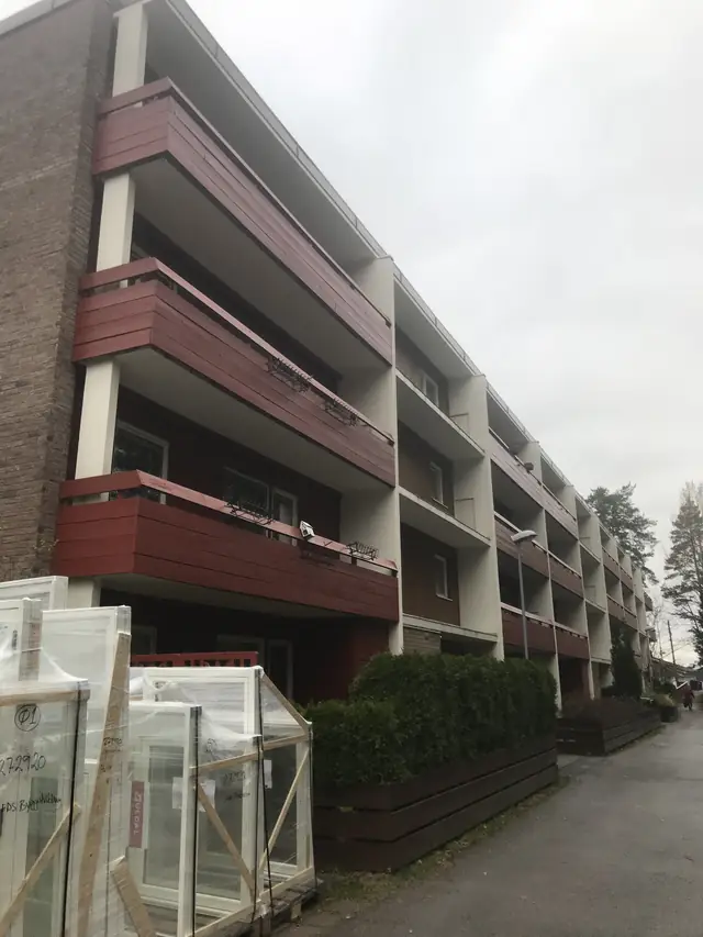 Boligblokk med gavlvegg i tegl, hvite søyler og røde balkongfronter. Nederst til venstre står paller med innpakkede hvite vinduer.